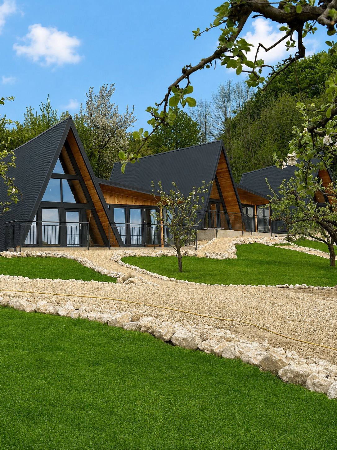 Trei cabane A-frame negre, așezate în șir pe un deal verde, cu pădurea în spate.